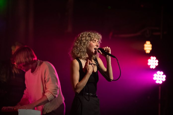 Stage lights illuminate Alaina Moore, who is singing with her eyes closed. Husband and musical partner Patrick Riley is bent over his keyboards to the left.