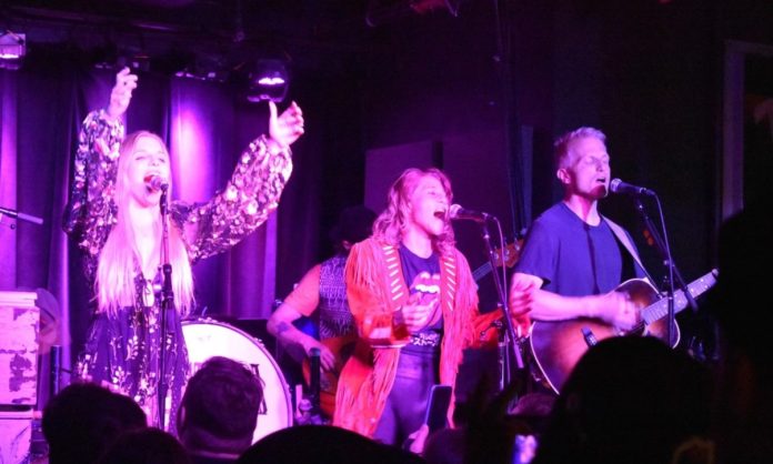 Three singers on stage with eyes closed. The singer on the left has her arms raised, the one in the center is dancing, and the one on the right is playing guitar.