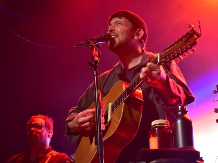 A bassist looks on in the background beside a singer/guitarist who is closing his eyes while singing into a microphone.