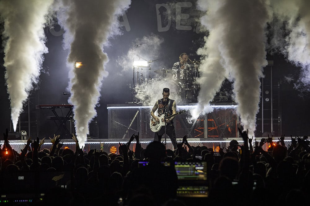 guitarist on stage in front of drummer with several smoke plumes firing upwards